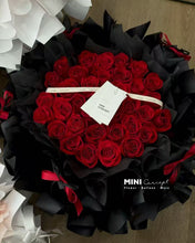 Load image into Gallery viewer, Heart-shaped bouquet of red roses with black ribbons and a visible brand tag on a wooden surface.

